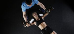 Female athlete performing dumbbell exercises with trainer's guidance in a gym setting.
